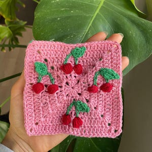 Puede incluir: Un cuadrado rosa tejido a ganchillo con un diseño de cerezas. El cuadrado presenta un patrón repetido de cerezas rojas con hojas verdes. El artículo hecho a mano es probablemente un posavasos o una pieza decorativa.