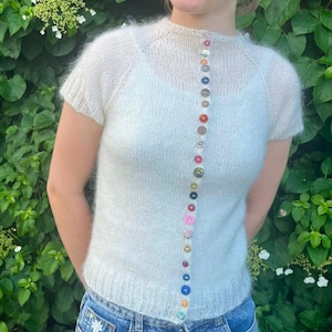 Può includere: Un maglione in maglia color bianco sporco a maniche corte, caratterizzato da una consistenza soffice e uno scollo rotondo. Una fila verticale di piccoli bottoni multicolori corre lungo il centro del capo. Il maglione ha orlo e polsini a coste.