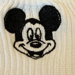 May include: Close-up of a light blue knit hat featuring a black and white embroidered Mickey Mouse face. The hat has a ribbed texture and a classic cartoon character design.