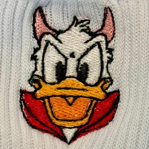 May include: A light blue knit fabric featuring an embroidered patch of a white duck character with a yellow beak, black eyes, pink devil horns, and a red collar. The embroidery uses detailed stitching to define the facial features and colors.