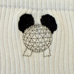 May include: Close-up of a white knit fabric with a panda face design. The panda has black embroidered ears and a silver, textured face. The knit fabric has vertical ribbing.