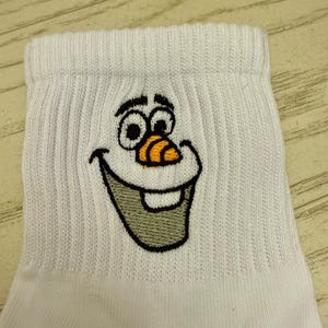 May include: White socks with an embroidered Olaf face from the movie Frozen. The Olaf face features black eyes, a carrot nose, and a smiling mouth. The socks are made of a ribbed knit fabric.