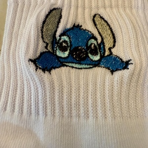 May include: White ribbed socks featuring an embroidered image of the blue alien character Stitch from the animated film Lilo & Stitch. The character has large eyes, a black nose, and gray ears. The socks are made of a soft, knitted material.