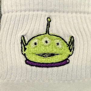May include: Close-up of a light blue ribbed sock featuring an embroidered green alien character with three eyes, black outlines, and a purple collar. The alien has a smiling expression.