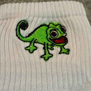 May include: Close-up of a white ribbed sock featuring a green embroidered chameleon design. The chameleon has black outlines, red mouth, and brown eyes. The sock is made of a soft, textured material.