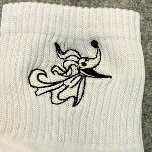 May include: A white ribbed sock featuring a black embroidered graphic of a ghost dog with long, flowing ears and a pointed nose. The embroidery is centered on the fabric, showcasing a detailed outline of the character against the white background.