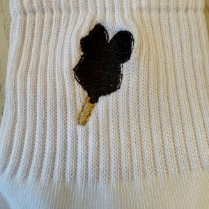 May include: White ribbed sock featuring a black ice cream bar design with a gold-colored stick. The sock has a folded cuff and is made of a soft, textured material. The ice cream bar design is embroidered.
