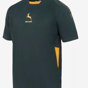 Puede incluir: Camiseta verde oscuro de manga corta con detalles dorados. Presenta un logotipo de impala dorado y el texto "SOUTH AFRICA" en el pecho. La camiseta tiene cuello amarillo y un panel lateral amarillo.