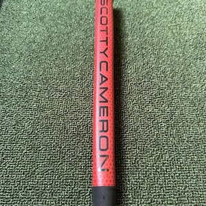 May include: A red golf club grip with the white text "SCOTTY CAMERON". The grip has a textured surface and a black base. The grip is on a green surface.