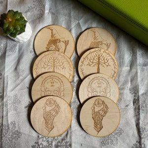 May include: Set of eight wooden coasters featuring various designs from The Lord of the Rings. The coasters are round and have a natural wood finish. The designs include the White Tree of Gondor, the One Ring, the Eye of Sauron, and the Fellowship of the Ring.