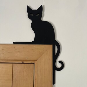 May include: Black silhouette of a cat sitting on a wooden door frame. The cat's tail curls around the frame.