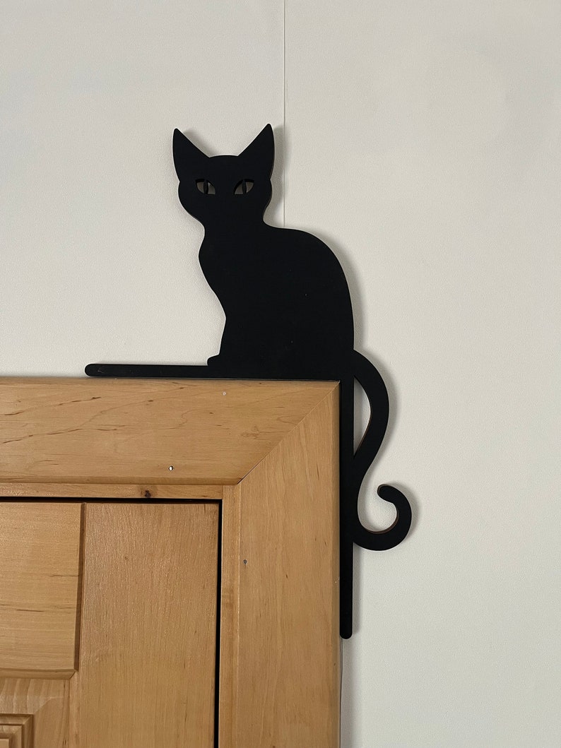 May include: Black silhouette of a cat sitting on a wooden door frame. The cat's tail curls around the frame.