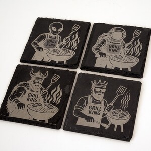 Puede incluir: Juego de cuatro posavasos cuadrados de pizarra negra con diseños grabados. Cada posavasos presenta un personaje diferente a la parrilla, incluyendo un alienígena, un astronauta, un vikingo y un rey. El texto "GRILL KING" está en cada posavasos.