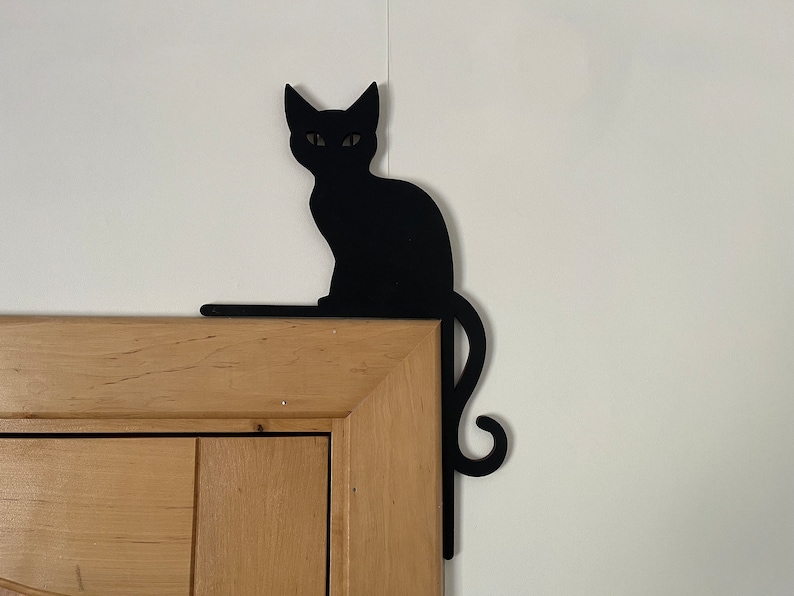 May include: Black metal silhouette of a cat sitting on a wooden door frame. The cat's tail curls around the frame.