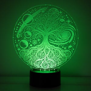 Può includere: Una luce notturna 3D stampata in verde illuminata con un design ad albero della vita. L'albero è circondato da pianeti, stelle e una galassia. La luce notturna è su una base nera.
