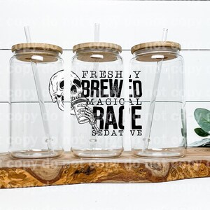 May include: Three clear glass tumblers with bamboo lids and straws, arranged on a wooden serving board. Each tumbler features a black and white graphic of a skull holding a coffee cup, with the words "FRESHLY BREWED MAGICAL RACE SEDATIVE".