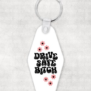 May include: White motel key chain with black text that reads "Drive Safe Bitch" in a retro font. Pink flowers are scattered around the text.