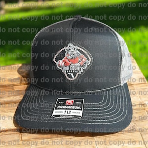 May include: Black trucker hat with a gray mesh back and a curved brim. The hat features a patch with a cartoon hog in a baseball uniform and the words "Hog County Baseball". The hat is adjustable and has the Richardson brand label.