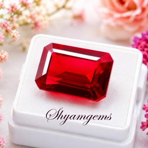 Puede incluir: Una gema roja de corte rectangular con bordes facetados, exhibida sobre un joyero cuadrado blanco. La caja presenta el texto Shyamgems en cursiva negra. La piedra muestra tonos carmesí profundos y una gran claridad visual.