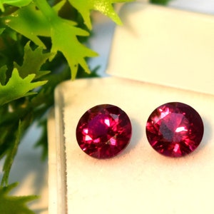 Puede incluir: Dos piedras preciosas redondas de color rojo oscuro en una caja blanca. Las piedras preciosas son facetadas y tienen un acabado brillante.