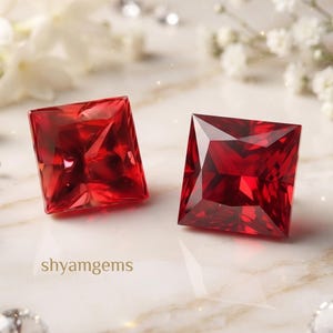 May include: Two square-cut, deep red gemstones with faceted surfaces reflecting light. The text shyamgems is printed in small brown letters near the stones. Several small, clear, round-cut diamonds and white flowers surround the red gems.