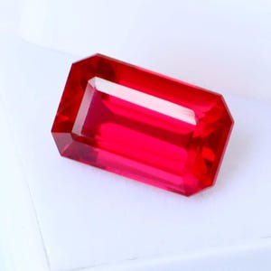 Peut inclure: Une pierre précieuse rubis rectangulaire et facettée, d'un rouge vif. La gemme est présentée dans un écrin en plastique blanc ouvert, mettant en valeur sa clarté et son éclat. La taille de la gemme présente de multiples facettes.