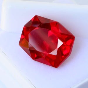 May include: A faceted, octagonal, deep red gemstone rests in an open white plastic display case. The gem has multiple reflective surfaces, catching the light. The case is on a white surface.
