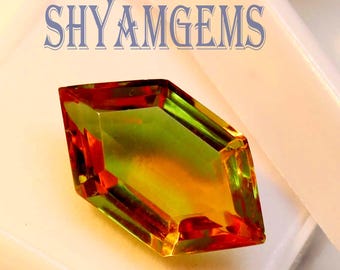 May include: A faceted gemstone with a mix of colors, including red, green, and yellow, is displayed in a white plastic case. The gemstone has a unique shape and is the focal point of the image. The text "SHYAMGEMS" is visible in the background.