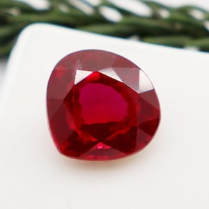 May include: A deep red gemstone with a faceted cut. The gemstone is set on a white background.