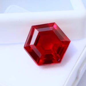 May include: A faceted, hexagonal, deep red gemstone. The gem is displayed in a white, open-top box, set against a white background. The gem's facets reflect light, creating a brilliant appearance. The overall theme is jewelry and gemstones.