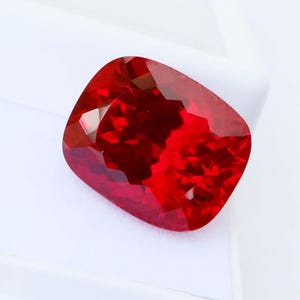 Può includere: Una gemma rosso intenso, taglio a cuscino. La gemma è posta su uno sfondo bianco, evidenziando le sue superfici brillanti e riflettenti. Il colore ricco e il taglio della pietra creano un effetto visivo sorprendente, suggerendo un gioiello di alta qualità.