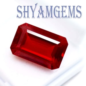 Pode incluir: Uma gema vermelha vibrante, de corte retangular, exibida numa caixa branca. A gema tem uma superfície polida e bordas afiadas, que refletem a luz. O texto "SHYAMGEMS" é visível acima da gema. Esta imagem mostra uma pedra preciosa, provavelmente para joalharia.