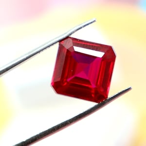 May include: A radiant, square-cut ruby gemstone held by silver tweezers. The ruby displays a deep red colour with a faceted surface, reflecting light. The background is a soft, blurred mix of colours, highlighting the gemstone's brilliance.