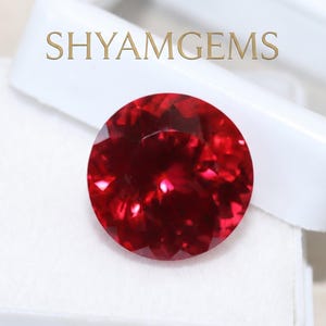 May include: A round, faceted, deep red gemstone with a brilliant cut, displayed on a white plastic jewelry box. The text SHYAMGEMS is printed in gold above the gemstone. The stone exhibits intense red color saturation and light reflection.