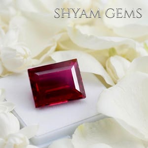 Könnte beinhalten: Ein rechteckiger, tiefroter facettierter Edelstein mit polierter Oberfläche. Der Stein liegt auf einem weißen Sockel, umgeben von weißen Blütenblättern. Der Text SHYAM GEMS ist in der oberen rechten Ecke des Bildes aufgedruckt.
