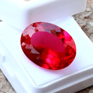 May include: A faceted, oval-shaped, vibrant red gemstone rests in a white display case. The gem's surface reflects light, showcasing its brilliant cut. The setting is simple, emphasizing the stone's color and clarity. This gemstone is likely for jewelry.