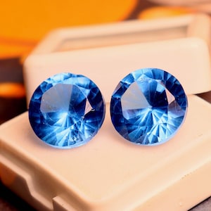 Può includere: Due cabochon di pietra preziosa blu in una scatola di gioielli bianca. Le pietre preziose sono rotonde e hanno un design sfaccettato.