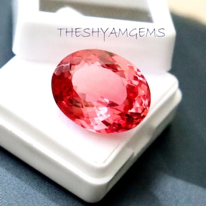 May include: A faceted, oval-shaped, pink gemstone rests in a white display box. The gemstone has a brilliant, reflective surface. The text "THESHYAMGEMS" is written in blue cursive above the gemstone.