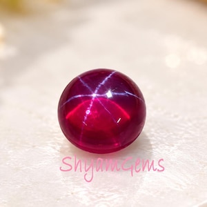 Pode incluir: Uma pedra preciosa de rubi estrela de formato redondo e corte cabochão, com uma cor vermelho magenta profundo. A pedra apresenta um efeito de estrela de seis raios distinto e centralizado, conhecido como asterismo, que aparece como um padrão.