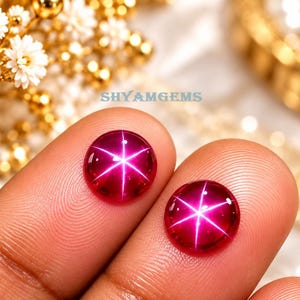 May include: Two round, cabochon-cut star rubies featuring a vibrant deep pinkish-red hue. Each gemstone displays a distinct, centered six-rayed star effect, known as asterism, which glows brightly against the smooth, polished surface of the stones.