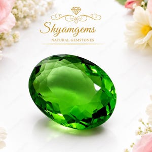 May include: A faceted oval green gemstone centered on a white background, surrounded by pink roses and white flowers. The text reads Shyamgems NATURAL GEMSTONES, Natural Gemstone, ETHICALLY SOURCED | 100% NATURAL | PREMIUM QUALITY.