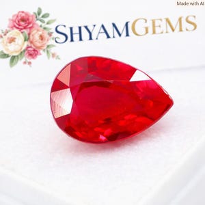 Peut inclure: Une pierre précieuse rouge taillée en forme de poire avec une finition brillante et translucide. La pierre est placée devant une carte blanche portant le texte SHYAMGEMS en lettres bleu foncé et une illustration décorative de roses roses et pêche.