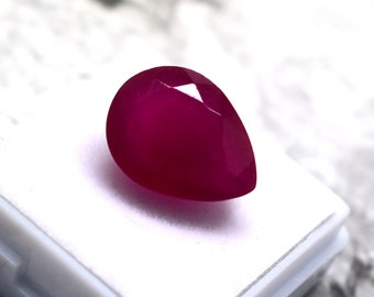 May include: A pear-shaped, faceted gemstone with a deep, saturated magenta-red colour. The stone features a smooth, polished surface with multiple geometric facets that reflect light, creating a brilliant appearance against a white display base.