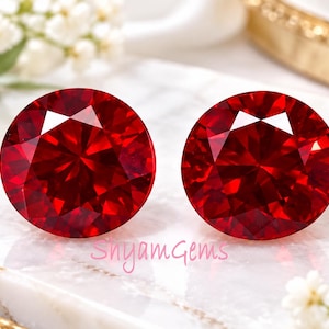 Può includere: Due pietre preziose rosse a taglio brillante rotondo con una superficie sfaccettata che riflette la luce. Le pietre sono di un colore cremisi vibrante e mostrano un motivo geometrico simmetrico. Il testo SnyamGems è stampato in rosa tra le pietre.