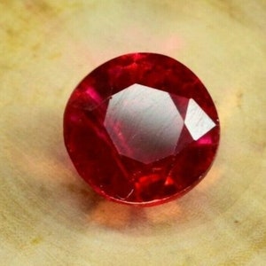 May include: A deep red gemstone with a faceted cut. The gemstone is round and has a high polish.