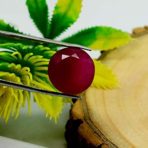 May include: A round, faceted, deep red gemstone held by silver-toned metal tweezers. The stone features a polished surface with geometric cuts that reflect light, showcasing a vibrant, saturated crimson hue throughout its circular, symmetrical form.