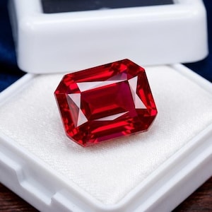 Puede incluir: Una gema roja rectangular, facetada y con corte brillante, exhibida dentro de un joyero blanco. La piedra preciosa muestra tonos carmesí profundos y bordes geométricos definidos, descansando sobre un inserto blanco de aspecto satinado.