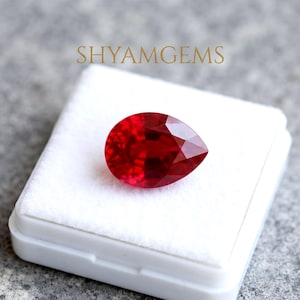 Könnte beinhalten: Ein birnenförmiger, facettierter roter Edelstein auf einem weißen Samtkissen. Der Schriftzug SHYAMGEMS ist in Gold über dem Edelstein aufgedruckt. Der Stein zeigt eine tiefe, leuchtende rote Farbe mit zahlreichen Facetten.