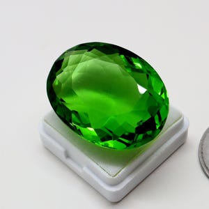 May include: A large, faceted, oval-shaped gemstone in a vibrant green hue. The gem is set on a white square base, showcasing its clarity and brilliance. The cut of the stone creates multiple reflective surfaces.