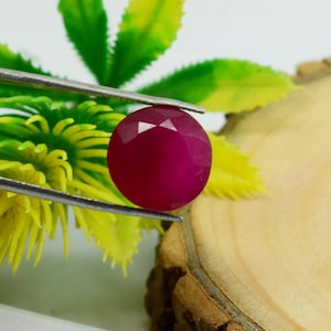 May include: A faceted, round, deep red gemstone is held by silver tweezers. The gem is a vibrant shade of red, with multiple facets catching the light. The background features green and yellow foliage and a wooden surface.